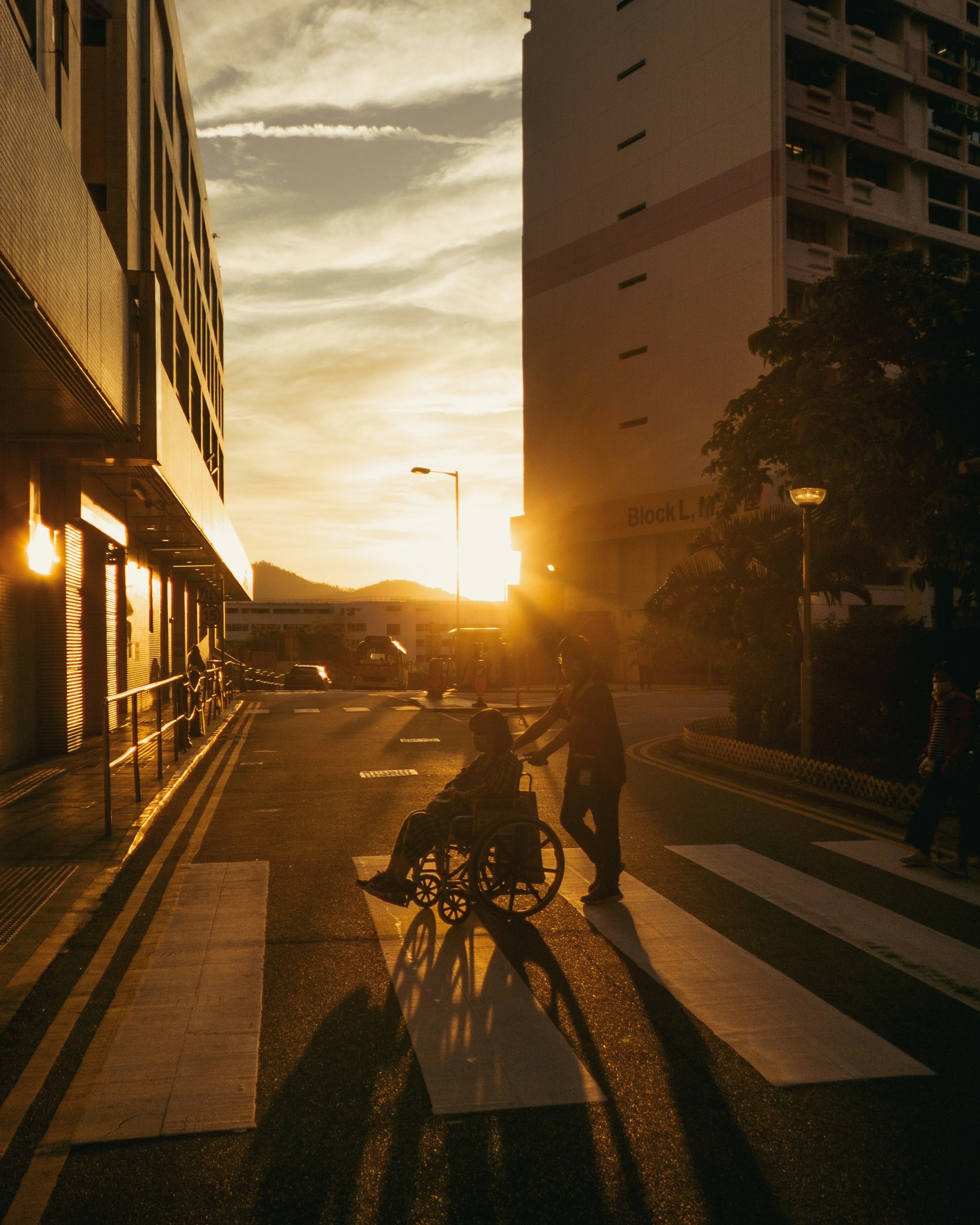 Someone pushes a person in a wheelchair across a city street at sunset.