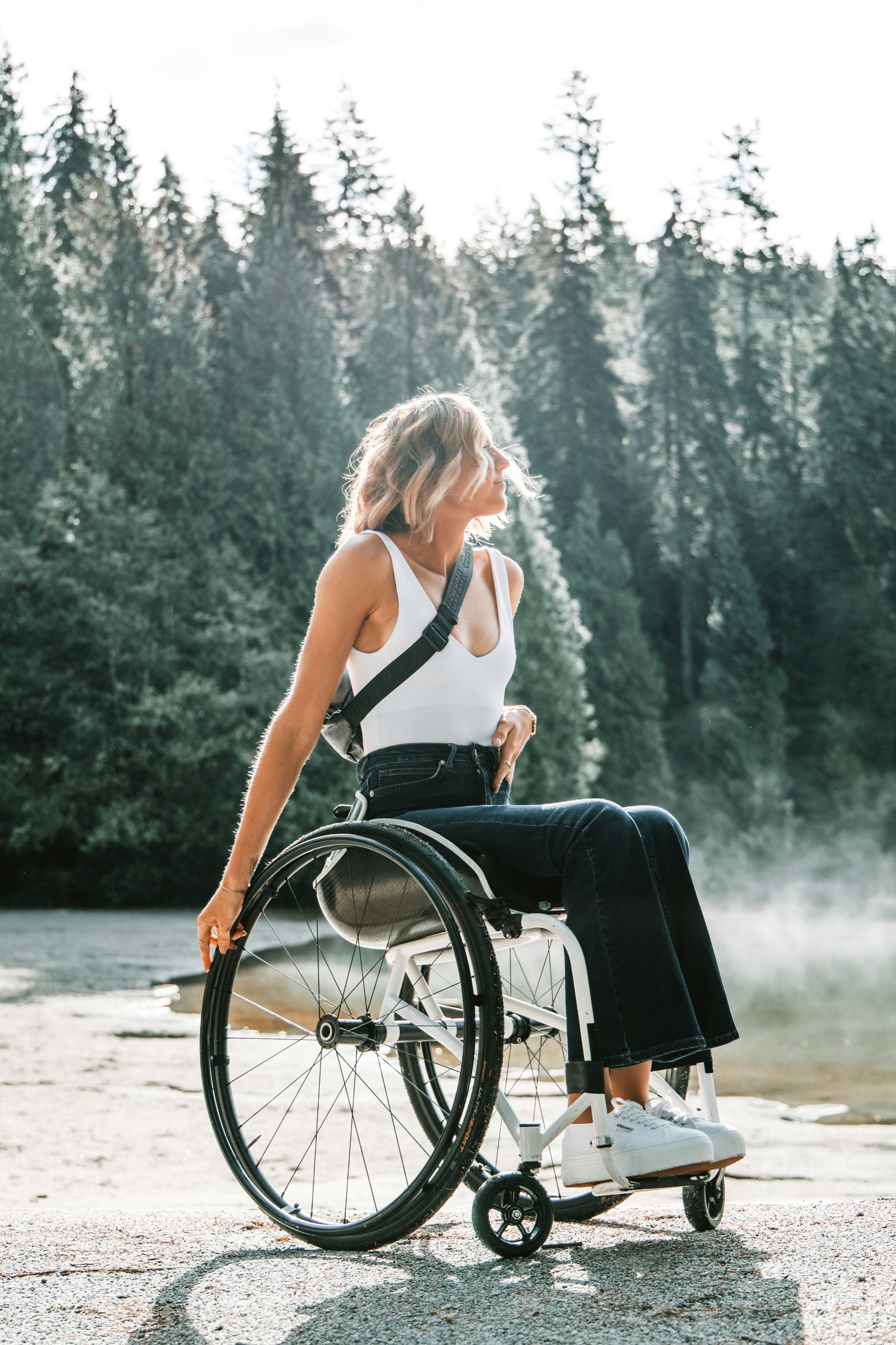 A young woman in a wheelchair by a lake, looking up at the trees in the morning sun.