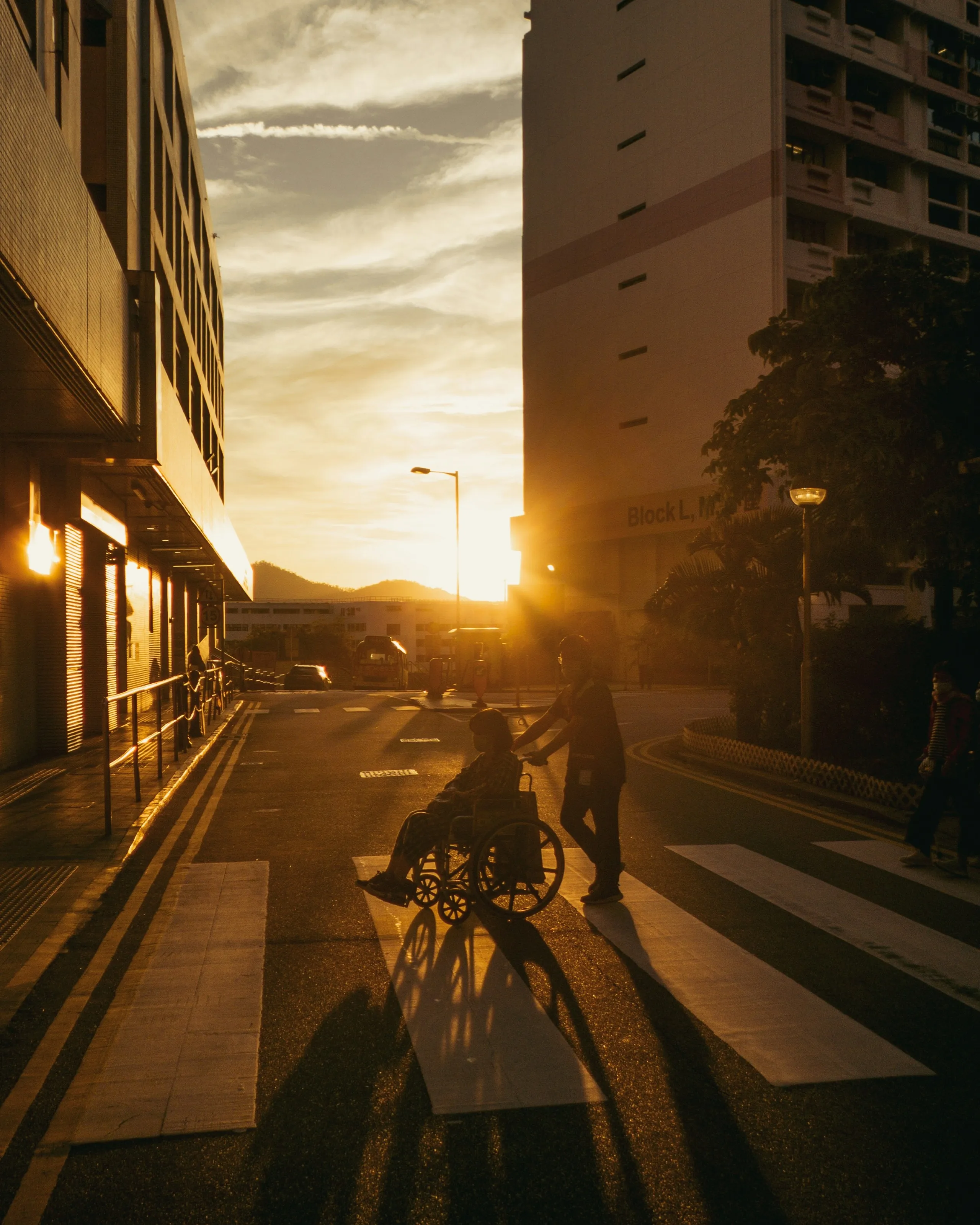 Someone pushes a person in a wheelchair across a city street at sunset.